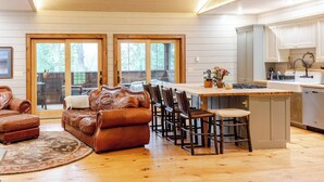 A spacious living room with a leather sofa and chairs, a wooden dining table, and a kitchen with white cabinets and a large island. - Secluded waterfront home, minutes from Lake Placid and Saranac Lake (Saranac Lake)