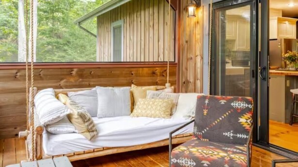 A cozy cabin interior with a wooden bed, a patterned chair, and a view of the outdoors. - Secluded waterfront home, minutes from Lake Placid and Saranac Lake (Saranac Lake)