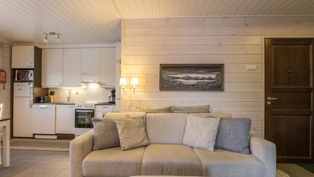 A living room with a sofa and ottoman, a kitchen in the background, and a wooden door. - Hil-lak-ka ii as 7 by Interhome (Äkäslompolo)