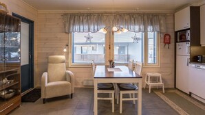A cozy dining area with a wooden table and chairs, a cushioned armchair, and a kitchen with a refrigerator and stove. - Hil-lak-ka ii as 7 by Interhome (Äkäslompolo)