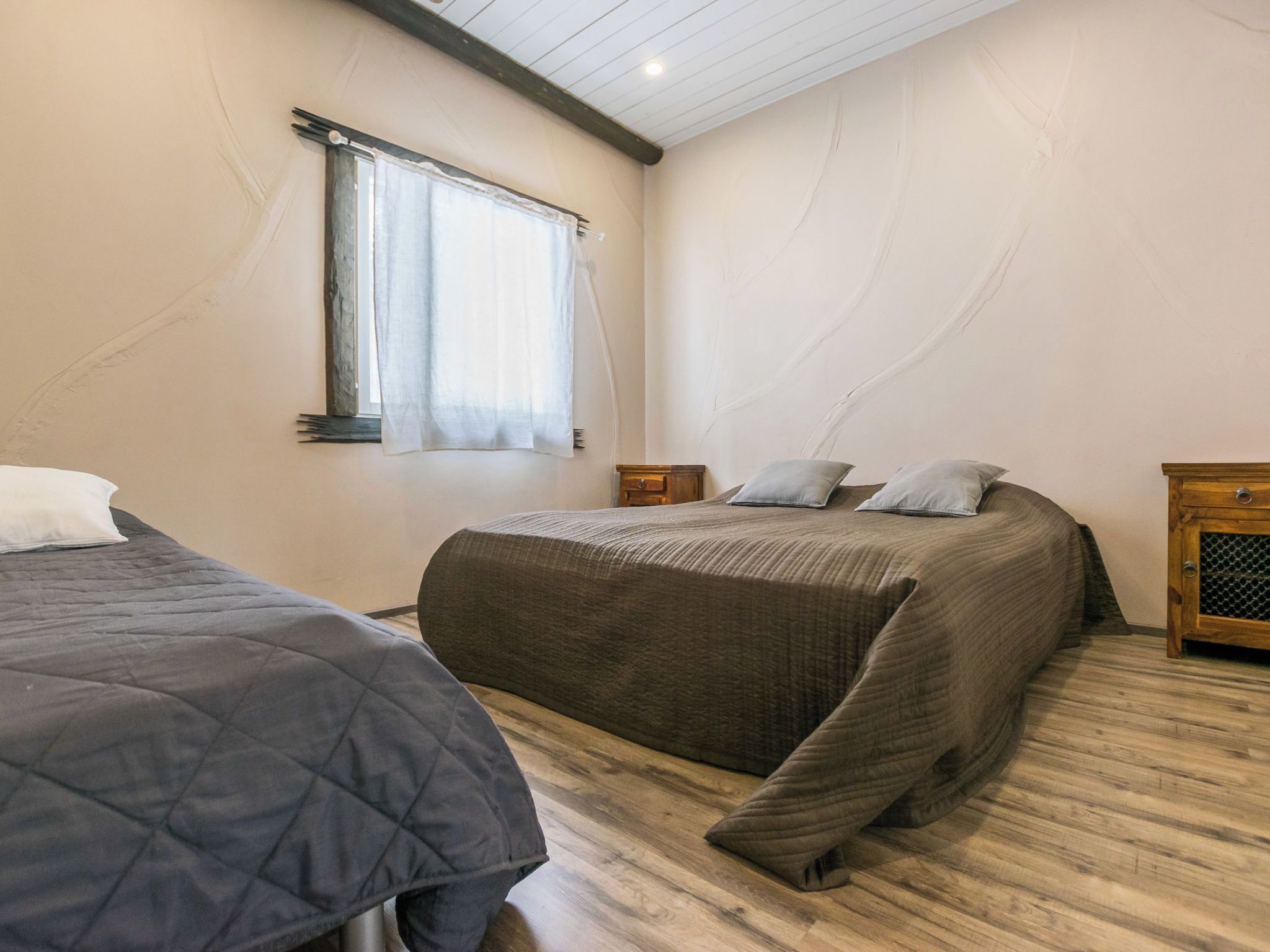A bedroom with two beds, a window with curtains, a wooden floor, and a nightstand with a lamp.