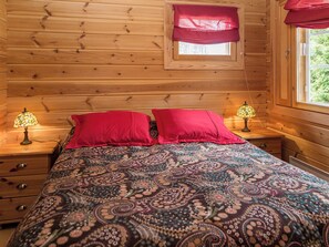 A wooden cabin bedroom with a patterned bedspread, two red pillows, two bedside lamps, and a window with a red valance. - Syvärinkulta b by Interhome (Nilsiä)