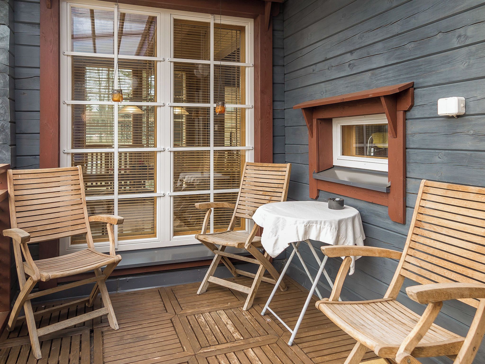 A wooden deck with four wooden chairs and a small round table, a window with blinds, and a wall-mounted light fixture.