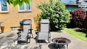 A patio with two lounge chairs, a small fire pit, and a brick house in the background.
