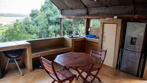 Une cuisine extérieure rustique équipée d’une cuisinière à gaz, d’un évier et d’un réfrigérateur. On y trouve une table et des chaises en bois, et la vue donne sur la forêt environnante.