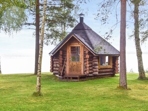A log cabin with a chimney, a door, and a window, surrounded by grass and trees.