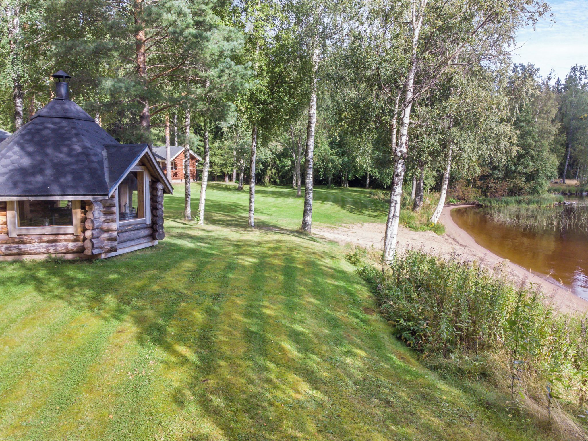 A log cabin with a chimney, surrounded by a grassy area and trees, near a body of water.