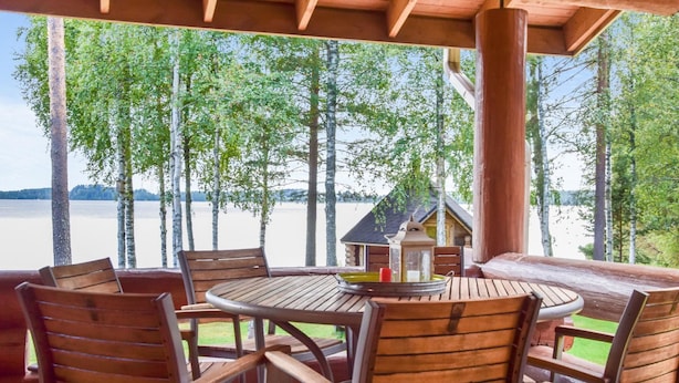 Une terrasse en bois avec une table ronde et des chaises, donnant sur un lac bordé d’arbres, avec un chalet visible au loin.