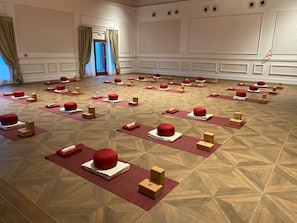 A spacious room with a patterned wooden floor, arranged seating with red cushions and wooden blocks, and a door with curtains.