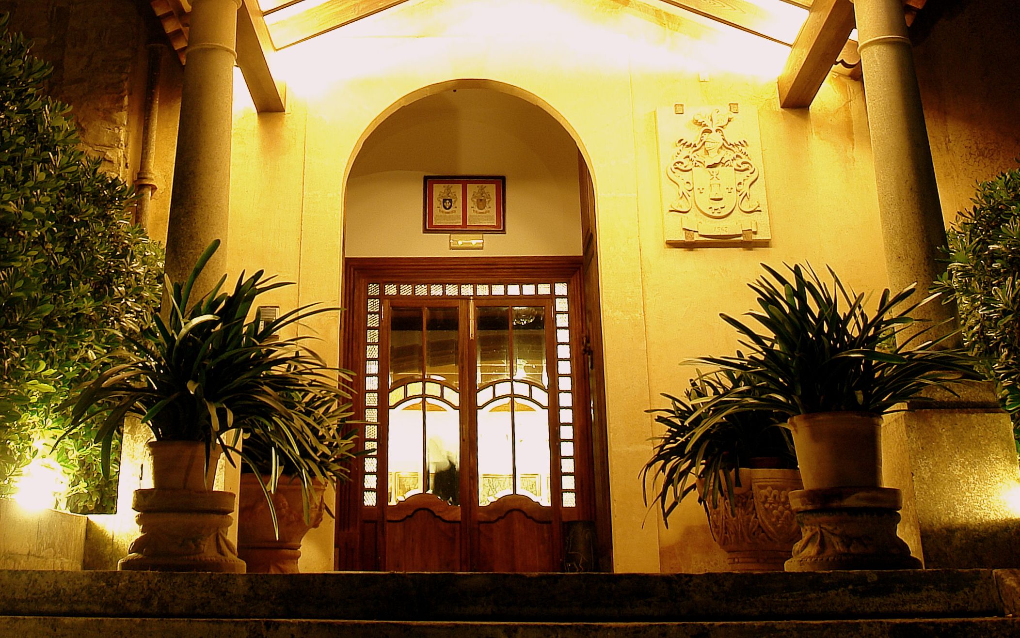 A building entrance with a wooden door, two potted plants, and a coat of arms plaque.
