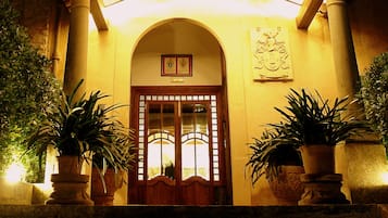 A building entrance with a wooden door, two potted plants, and a coat of arms plaque.