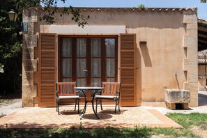 Une terrasse avec une table et des chaises, un banc en pierre et un bâtiment aux volets en bois et au toit en tuiles.