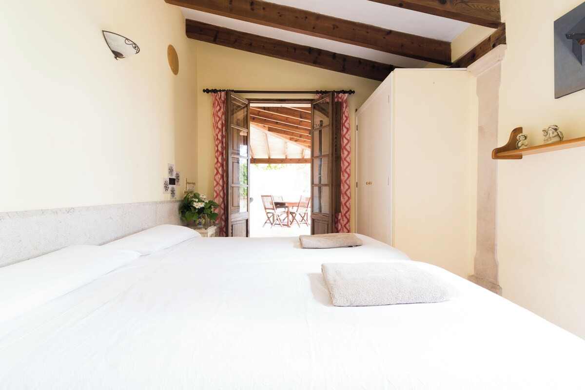 A hotel room with a large bed, a wooden ceiling, a small table with chairs, and a view through an open door.