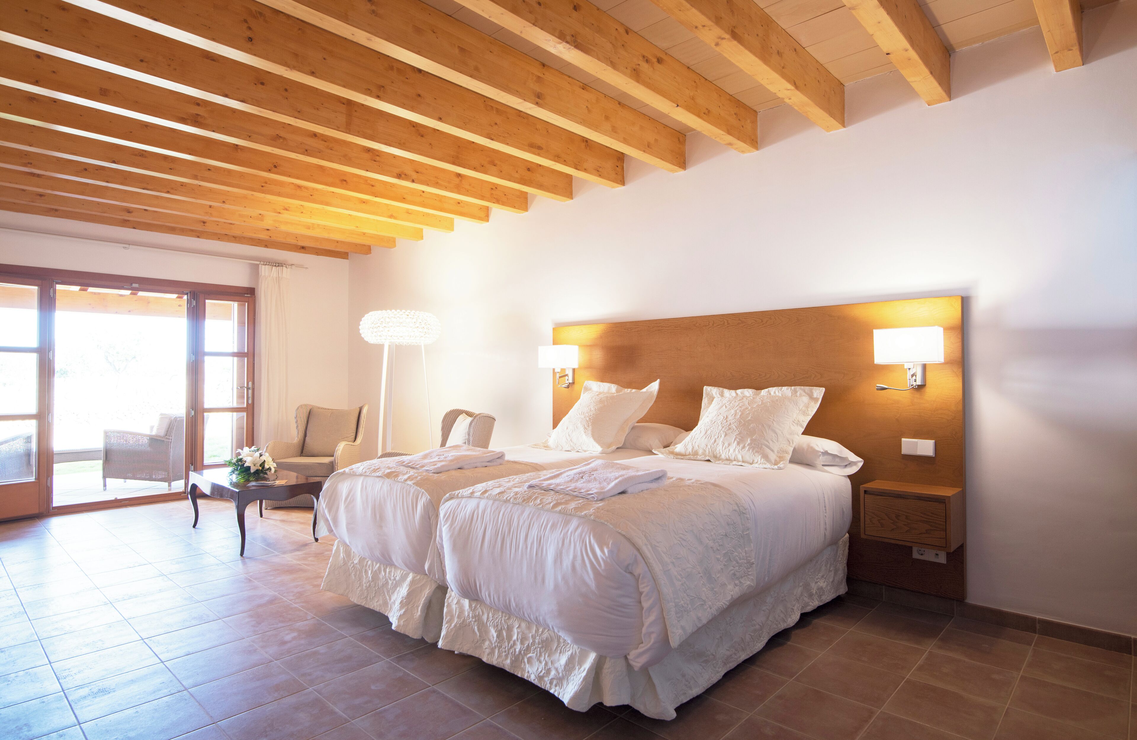 Een slaapkamer met twee bedden, houten balken in het plafond, een nachtkastje met een lamp en uitzicht door een glazen deur.
