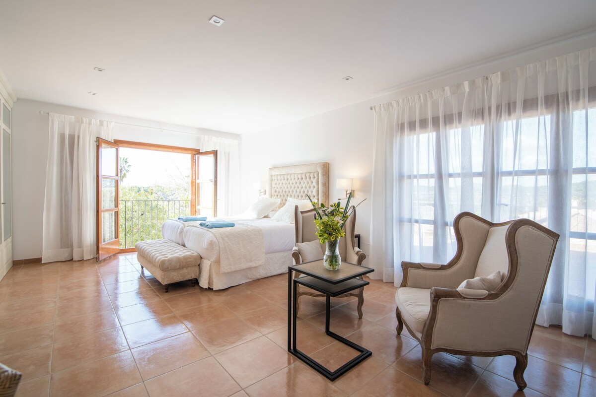 A spacious bedroom with a bed, a chair, a table, and a view of the outdoors.