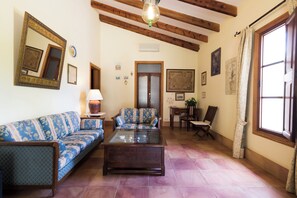 A living room with a sofa, a coffee table, and a chair. There is a door and a window with curtains. - Aumallia Hotel & Spa (Felanitx)