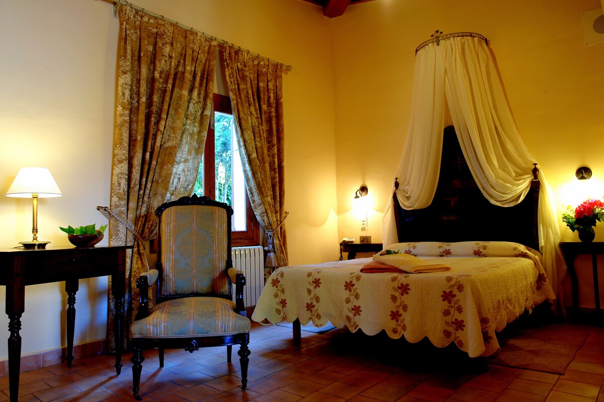 A bedroom with a four-poster bed, a chair, a table with a lamp, and a window with curtains.