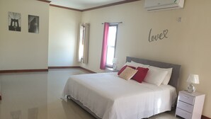 A spacious bedroom with a large bed, a nightstand, a wall-mounted air conditioner, and a window with curtains. - The Penthouse at Comfort Living (Kingston)