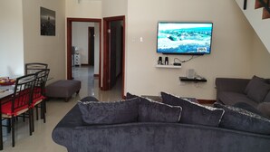 A living room with a sofa, a TV mounted on the wall, a dining area with a table and chairs, and a staircase. - The Penthouse at Comfort Living (Kingston)