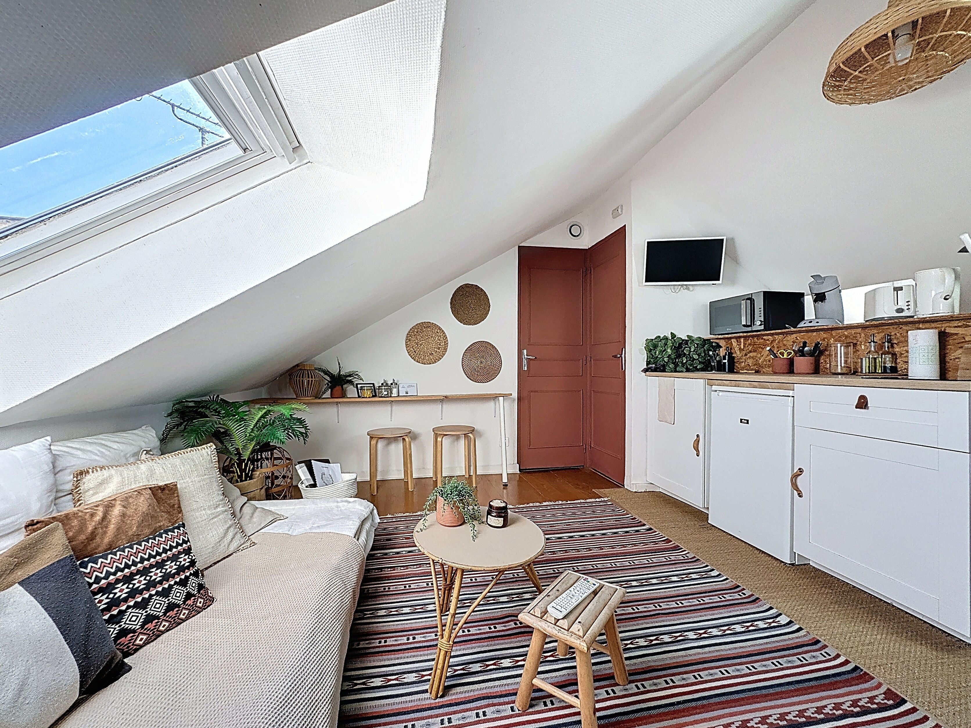 A cozy loft apartment with a high ceiling, a sloped ceiling with a skylight, a kitchenette, a dining area with stools, and a living space with a sofa and patterned rug.