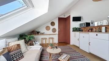A cozy loft apartment with a high ceiling, a sloped ceiling with a skylight, a kitchenette, a dining area with stools, and a living space with a sofa and patterned rug.
