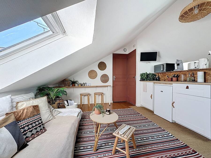 A cozy loft apartment with a high ceiling, a sloped ceiling with a skylight, a kitchenette, a dining area with stools, and a living space with a sofa and patterned rug.