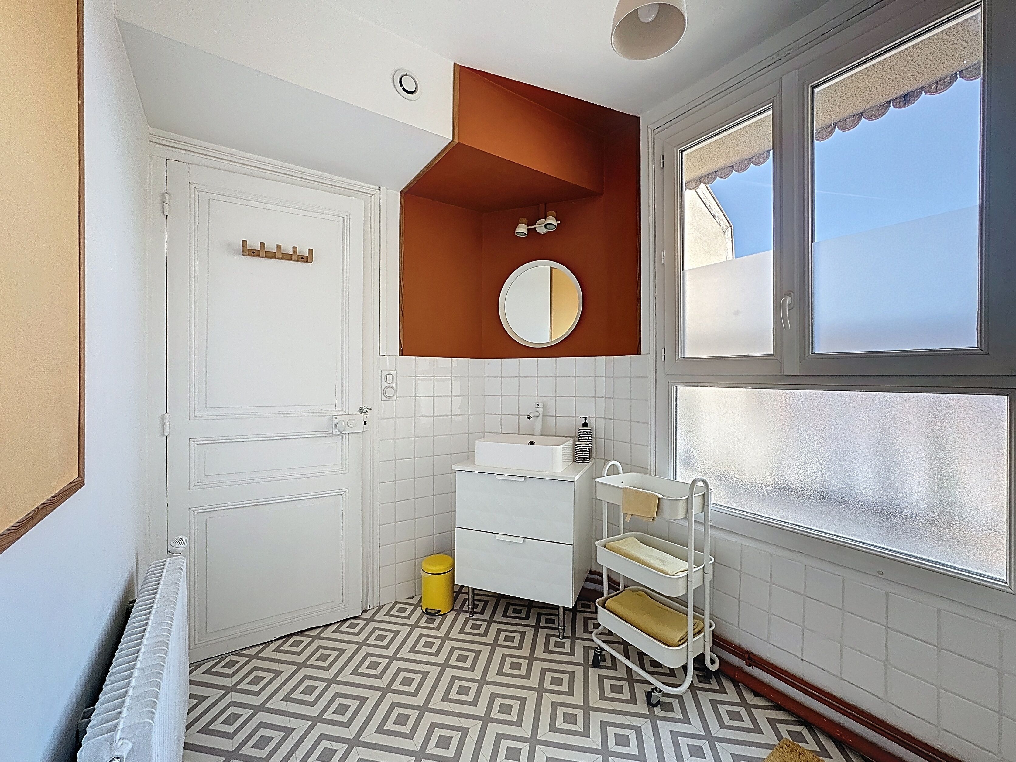 Un baño con puerta y lavamanos blancos, un espejo, un perchero para toallas y una ventana.