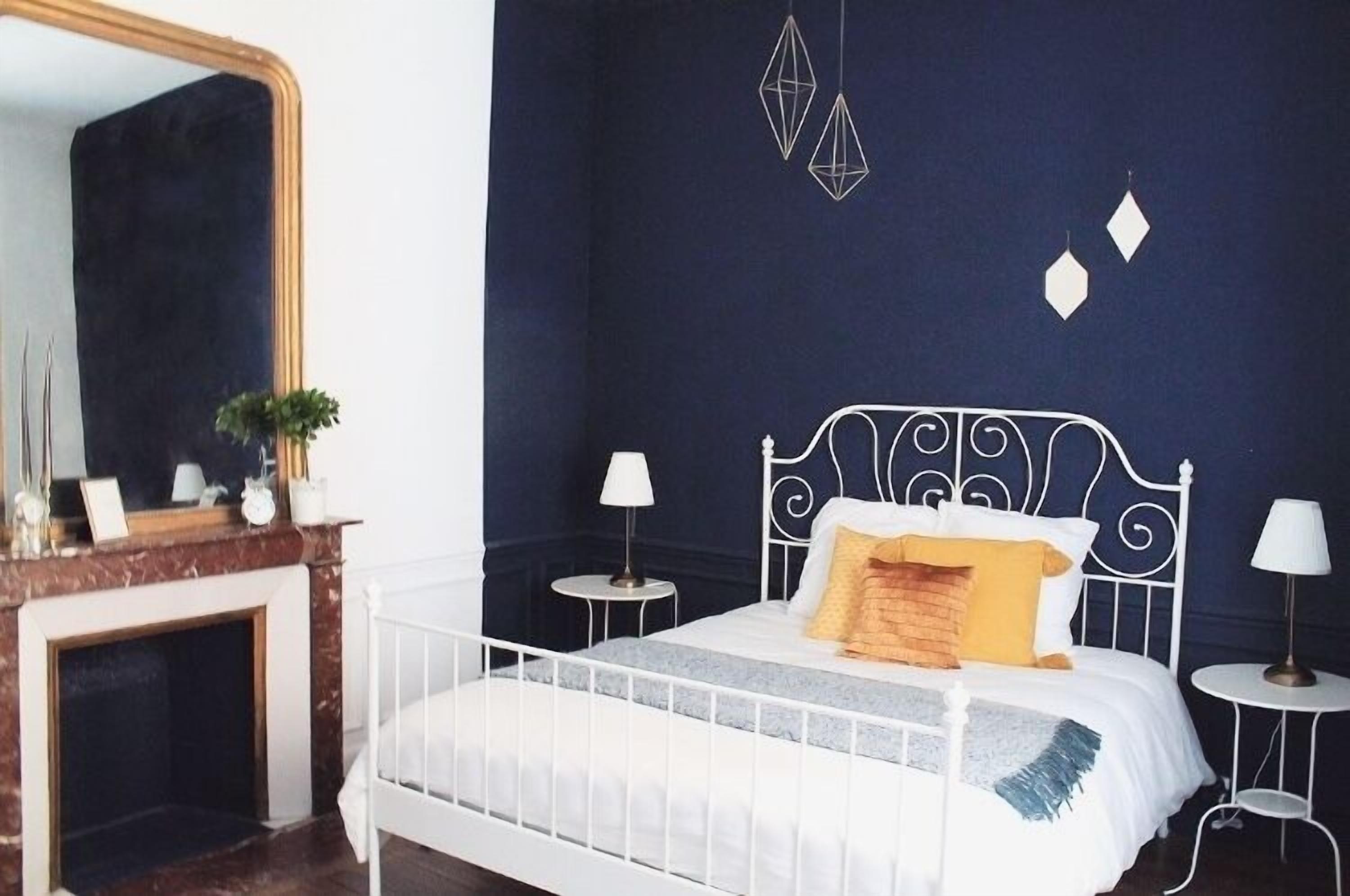 A bedroom with a white metal bed frame, a dark blue wall, a large mirror, and a small side table with a lamp.