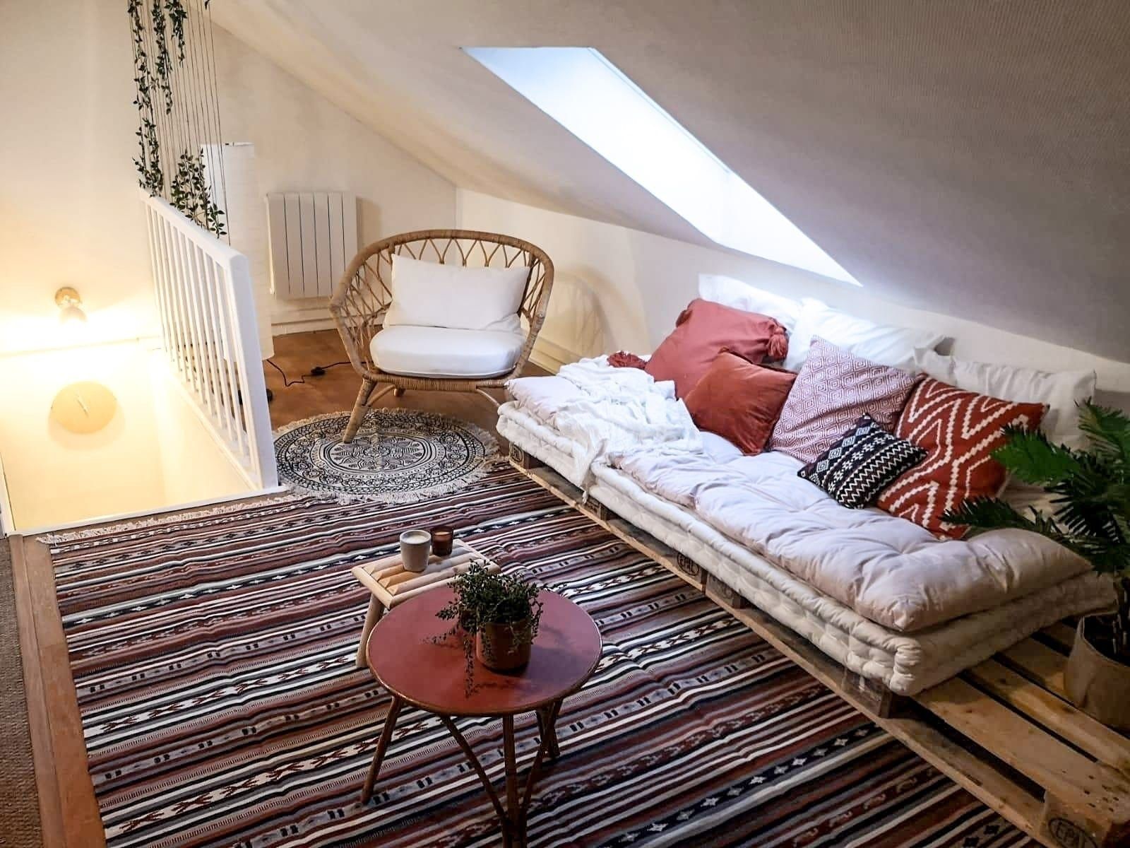 Une chambre à coucher confortable, aménagée en mezzanine, avec un plafond en pente, un fauteuil en rotin, un tapis à motifs, un canapé avec des coussins et une petite table sur laquelle est posée une plante.