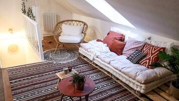 Een gezellig loft slaapkamer met een schuine kap, een rieten stoel, een kamerbreed tapijt met patroon, een bank met kussens en een kleine tafel met een plant.