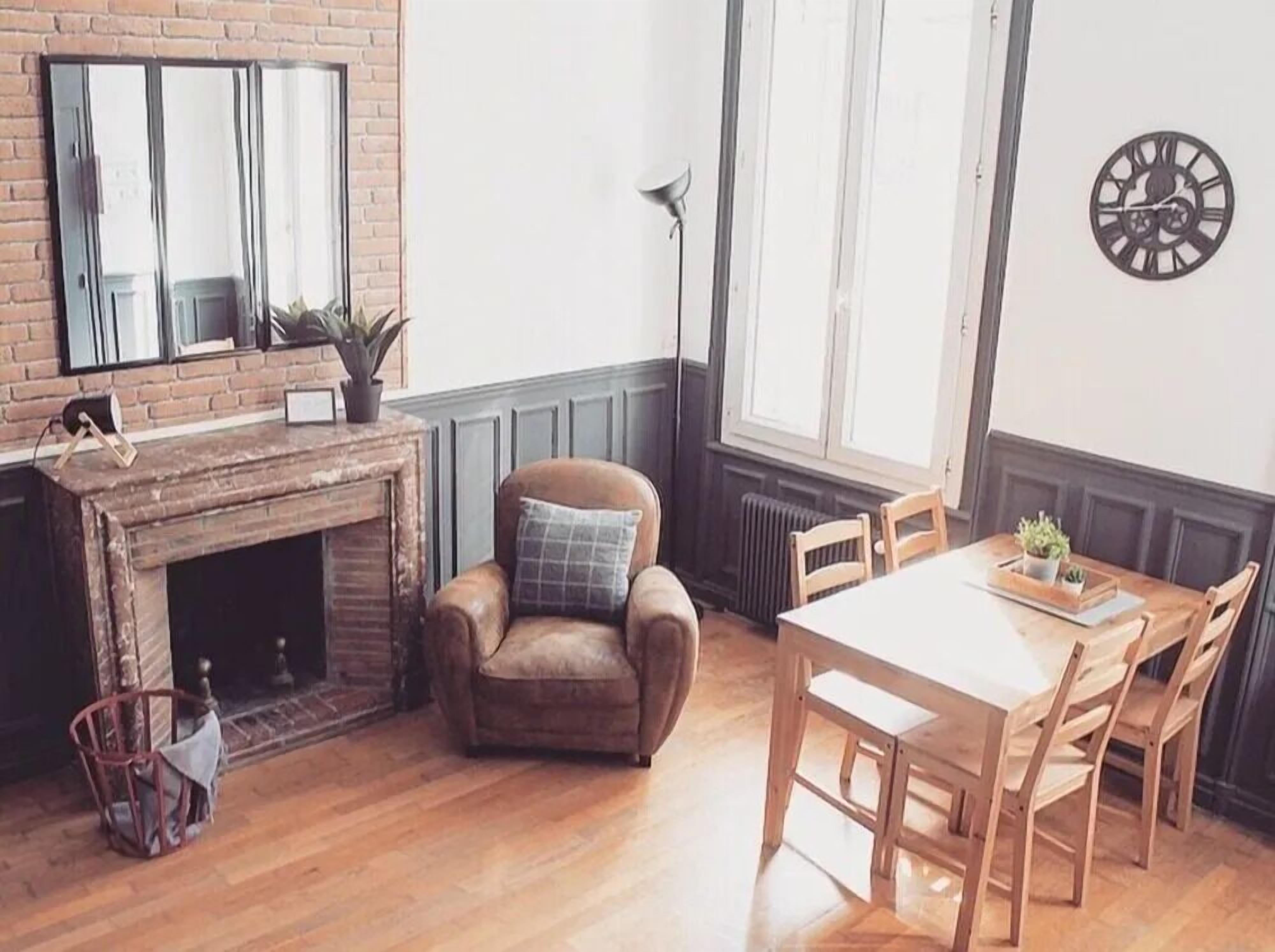 A cozy room with a brick fireplace, a leather armchair, a wooden dining table with chairs, and a large window with a view of the outside.
