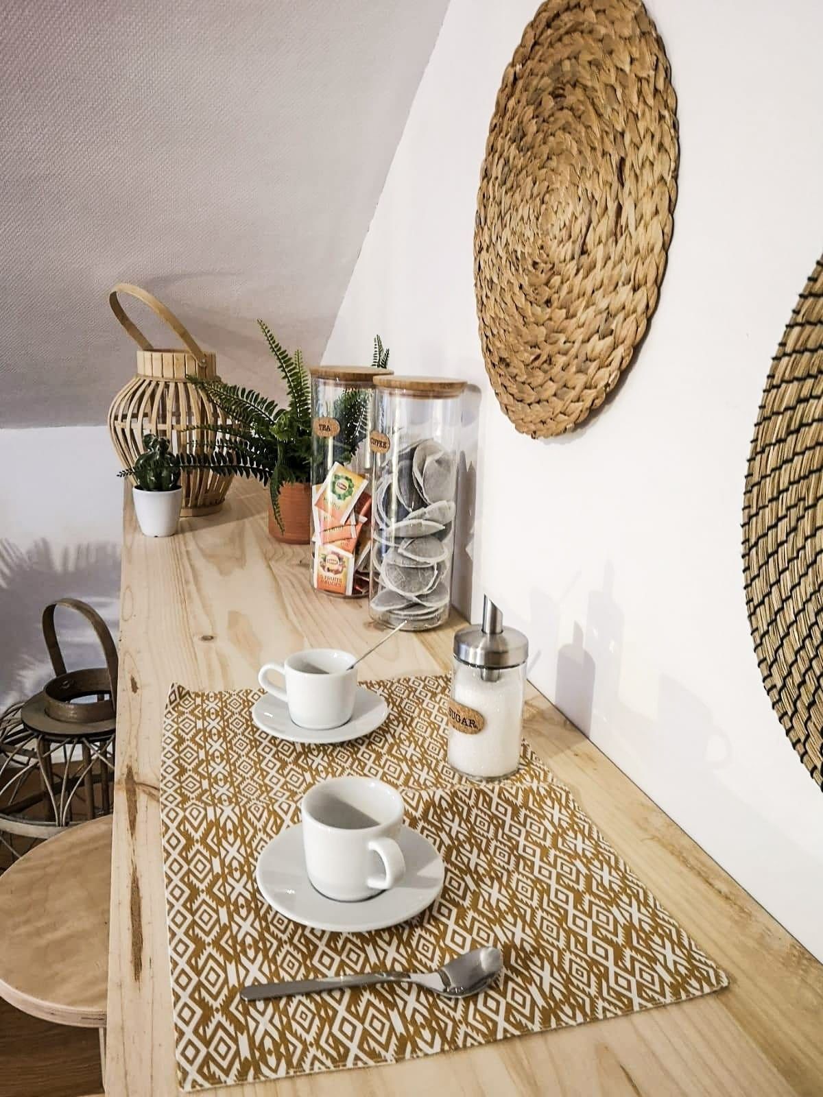 A wooden table with a woven placemat, two white cups, a sugar container, and a metal container with tea bags.