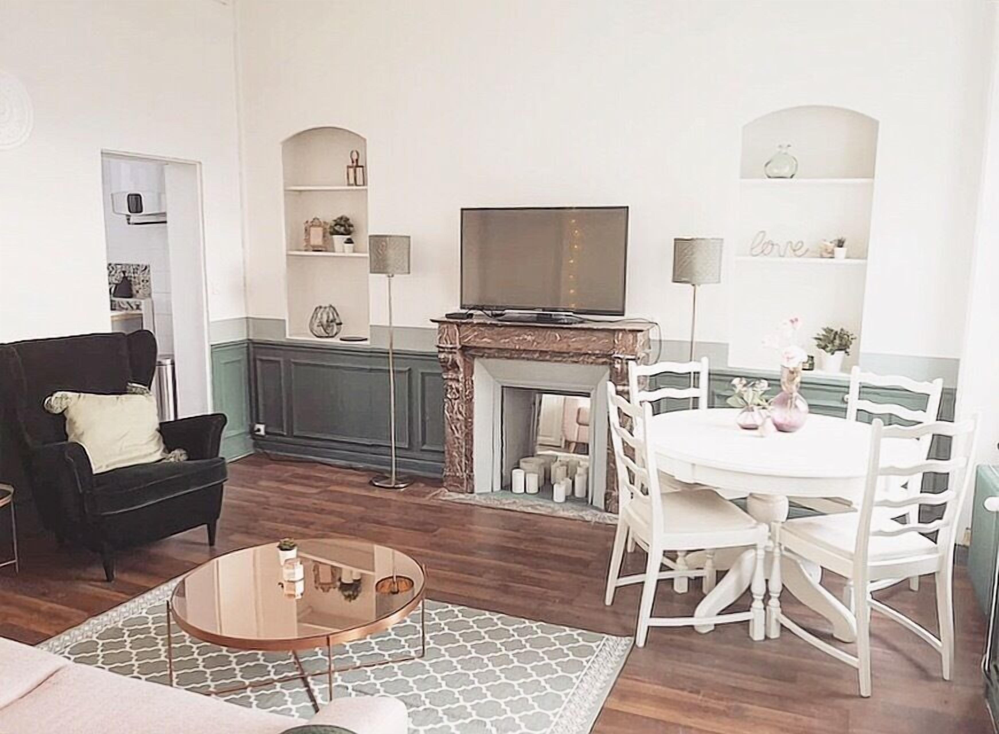 Un salon avec un canapé noir, une table basse ronde en verre, une table à manger blanche avec des chaises, une cheminée et un téléviseur posé sur un meuble.