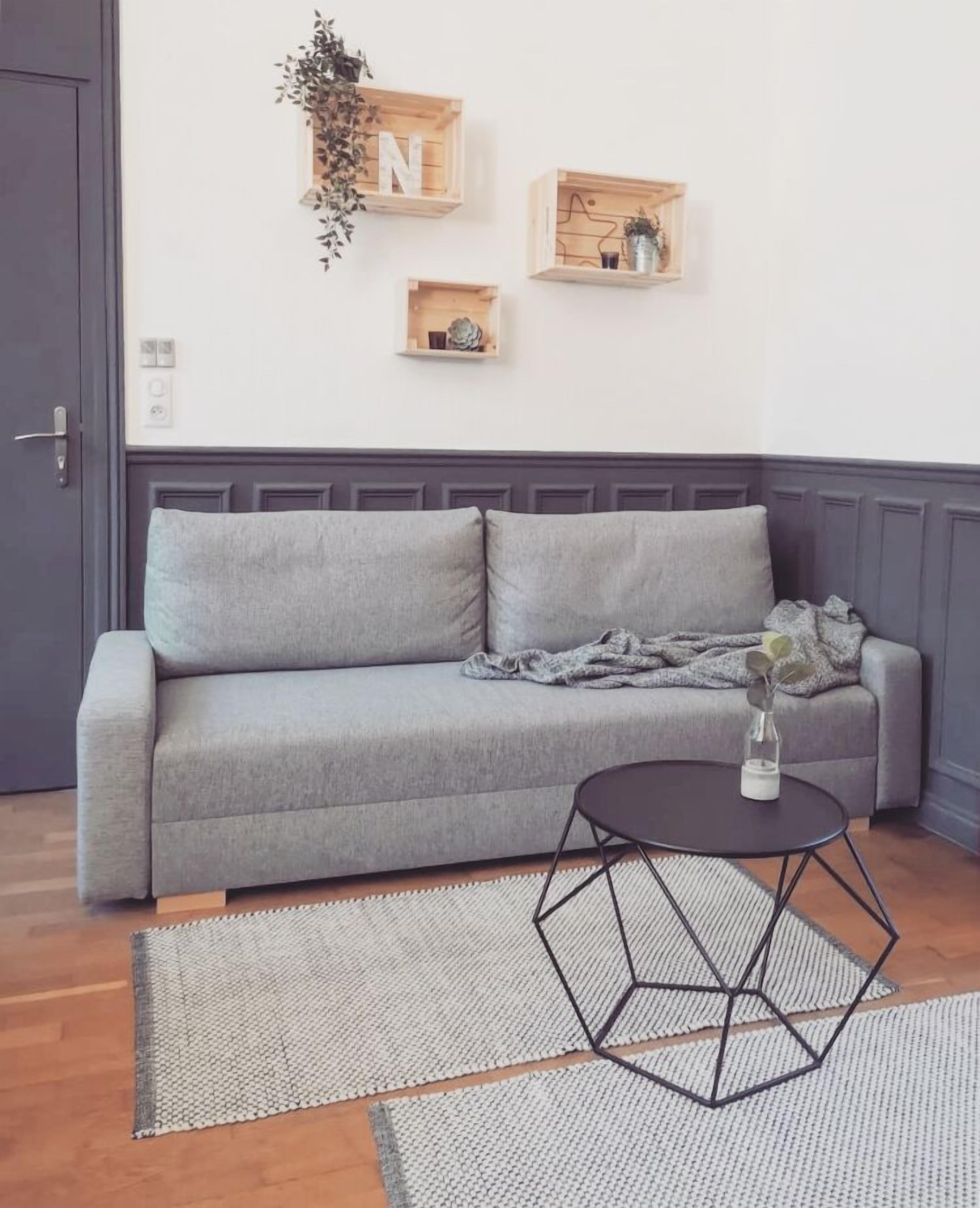Un sofá gris con una mesita de centro geométrica y una alfombra tejida en una habitación con estanterías de madera y una puerta.