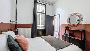 Une chambre à coucher avec un lit, un radiateur, une fenêtre avec des rideaux, une porte, un miroir et une étagère avec une plante.