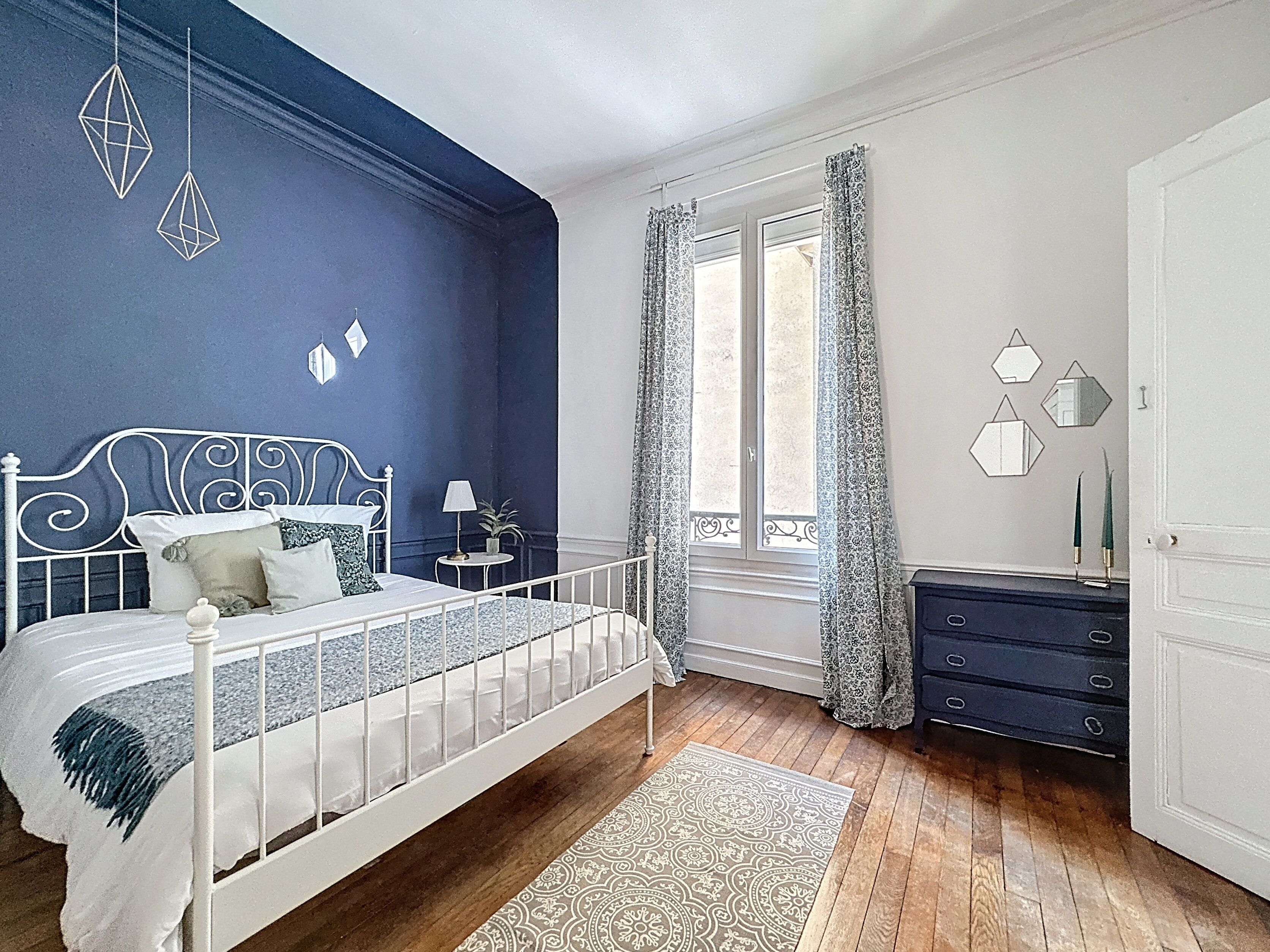 Un dormitorio con estructura de cama de metal blanco, pared de acento azul, ventana con cortinas estampadas y suelo de madera.