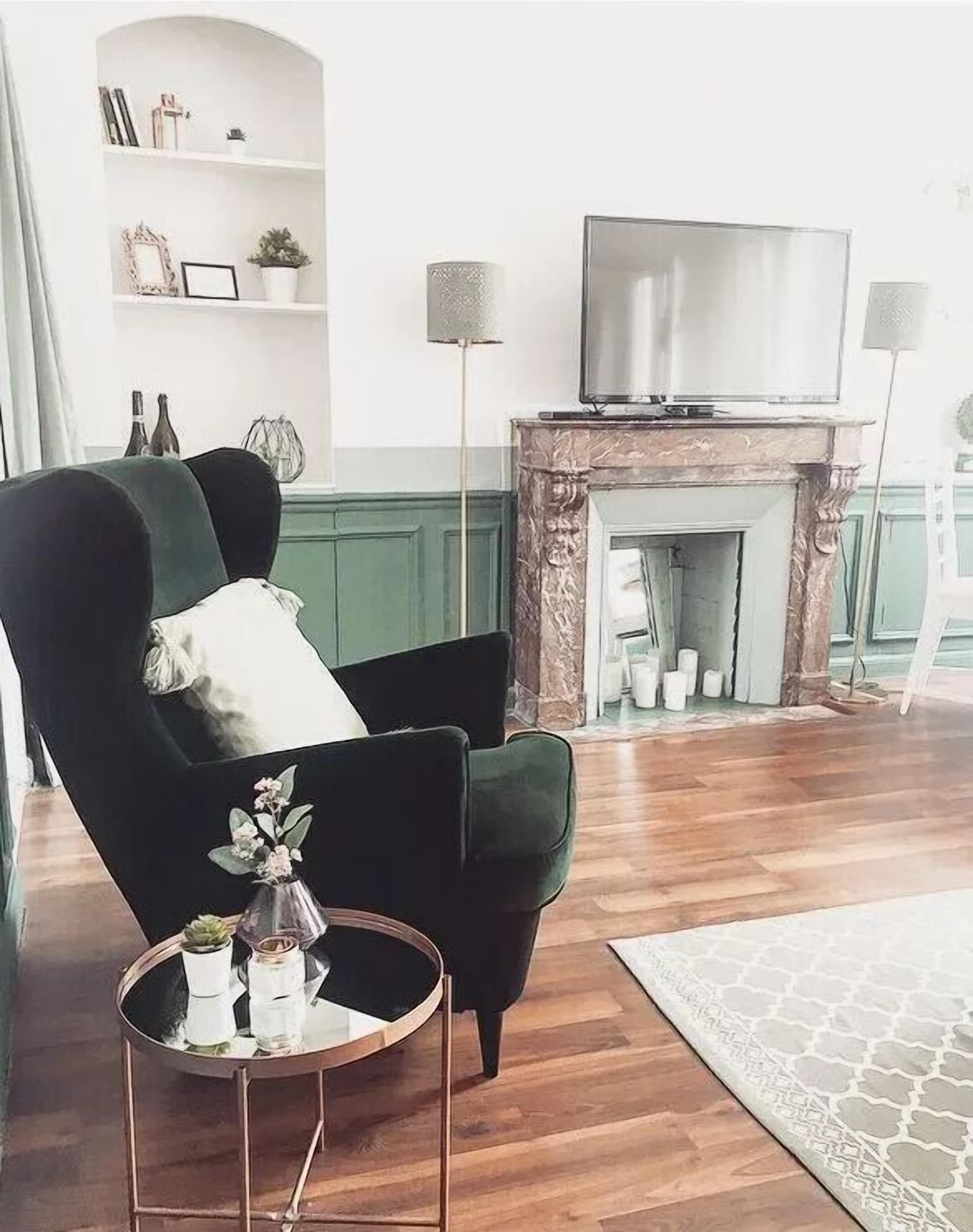 Un salon avec un fauteuil vert foncé, une table basse ronde ornée d’objets décoratifs, une cheminée et un téléviseur posé sur la manteau de la cheminée.