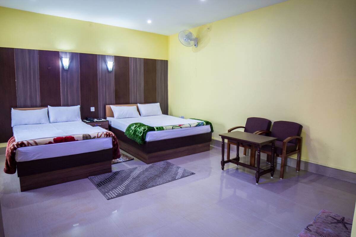 A hotel room with two beds, a small table, two chairs, a fan, and a rug.