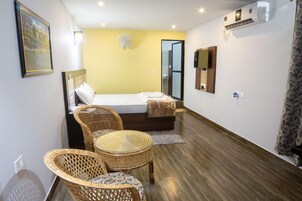 Una habitación de hotel con una cama, dos sillones de mimbre, una mesita, un ventilador, un equipo de aire acondicionado y un espejo en la pared.