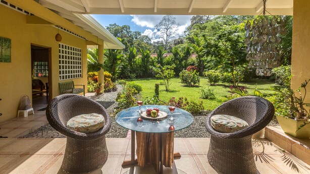 Une terrasse dotée de chaises en osier et d’une table en verre, aménagée pour deux personnes, et offrant une vue sur un jardin luxuriant, parsemé de différentes plantes, sous un ciel dégagé.