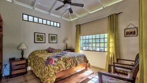 Une chambre à coucher comprenant un lit, une chaise, un ventilateur de plafond, une fenêtre avec des rideaux et un tableau accroché au mur.