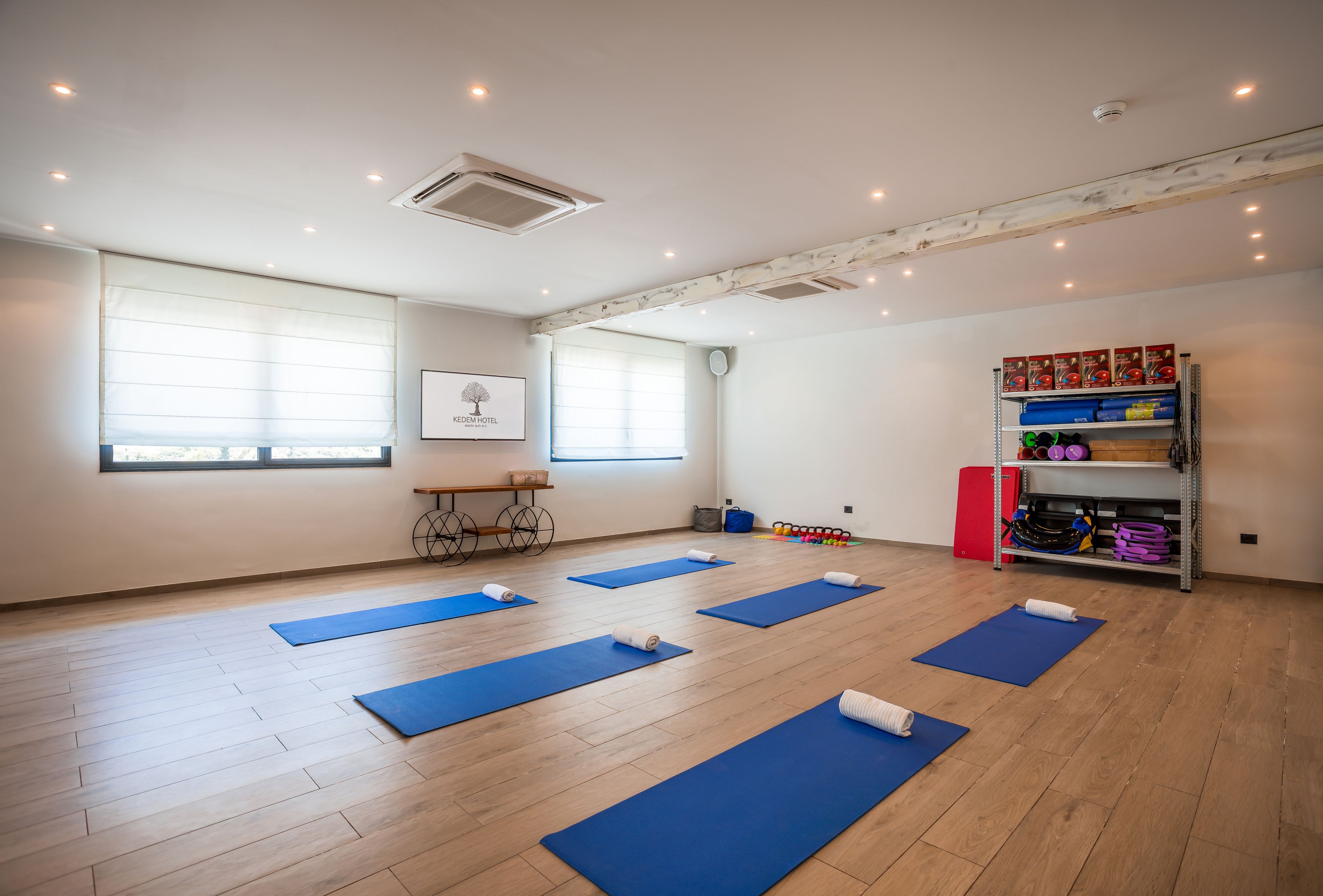 Una sala de yoga con colchonetas azules, suelo de madera, ventilador de techo y estante con equipo de ejercicio.