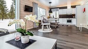 Una cocina moderna con armarios blancos, una zona de comedor con mesa y sillas blancas, y una sala de estar con sofá y una orquídea en maceta.