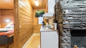 L’intérieur chaleureux d’un chalet avec des murs en bois, une cheminée en pierre, un coin cuisine et un lit recouvert d’une couverture rouge.