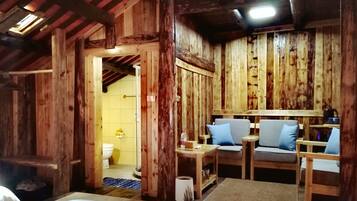 A rustic wooden room with a bed, a small table, and two chairs. There is a bathroom visible through an open doorway.