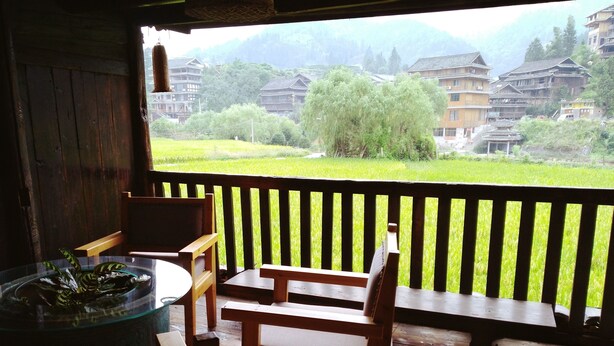 Una terraza de madera con una mesa y sillas, con vistas a un campo verde y edificios tradicionales al fondo.