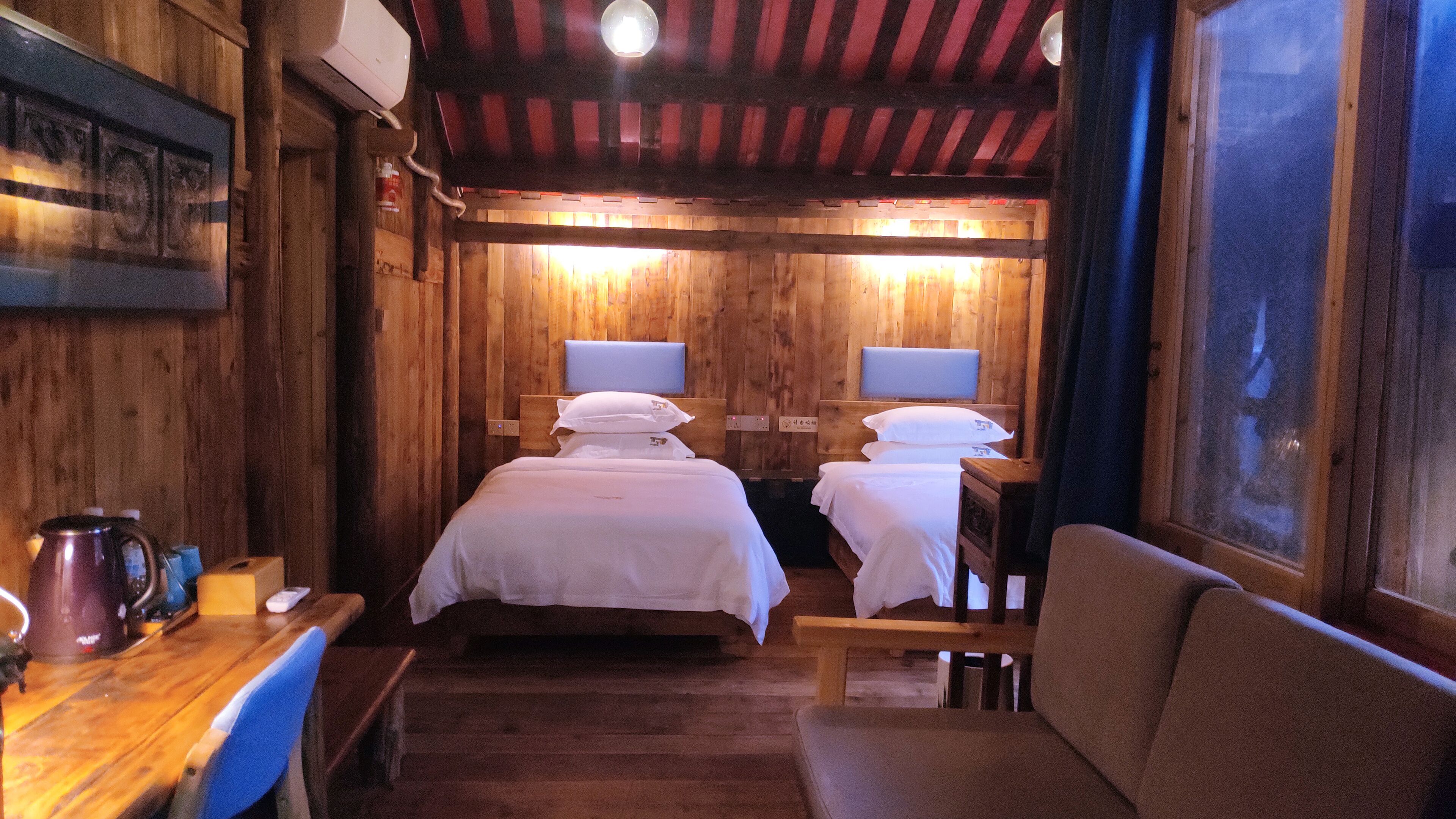 A cabin-style room with two beds, a wooden floor, a table with a kettle, and a window with a view.