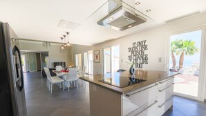 A modern kitchen with a large island, dining area with white chairs, and a view of palm trees outside. - LA CORNICHE D'ARGENT VI4136 by RIVIERA HOLIDAY HOM (Niza)