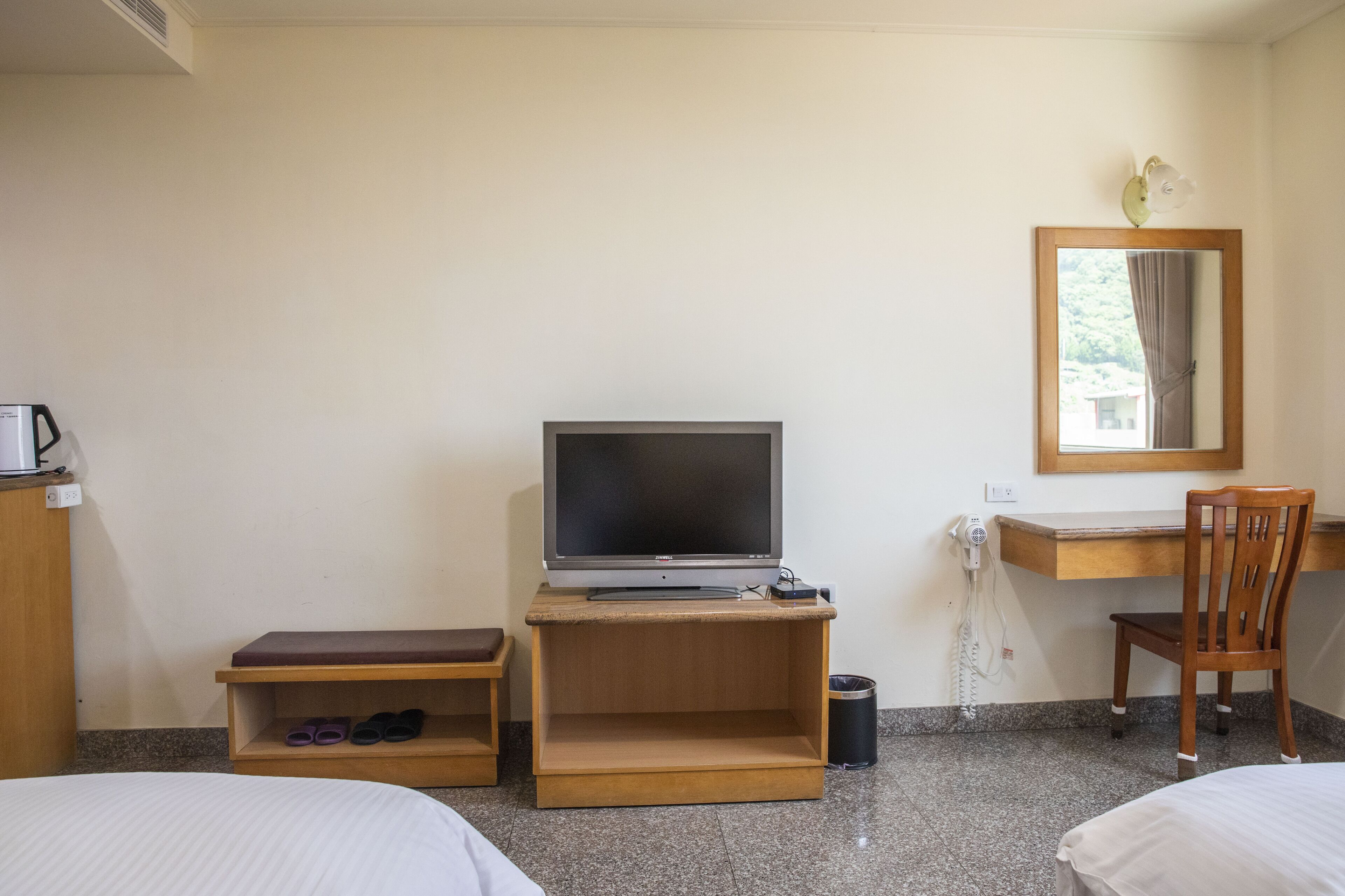 Habitación de hotel con cama, escritorio con computadora, silla, estante pequeño con zapatos y un cubo de basura.