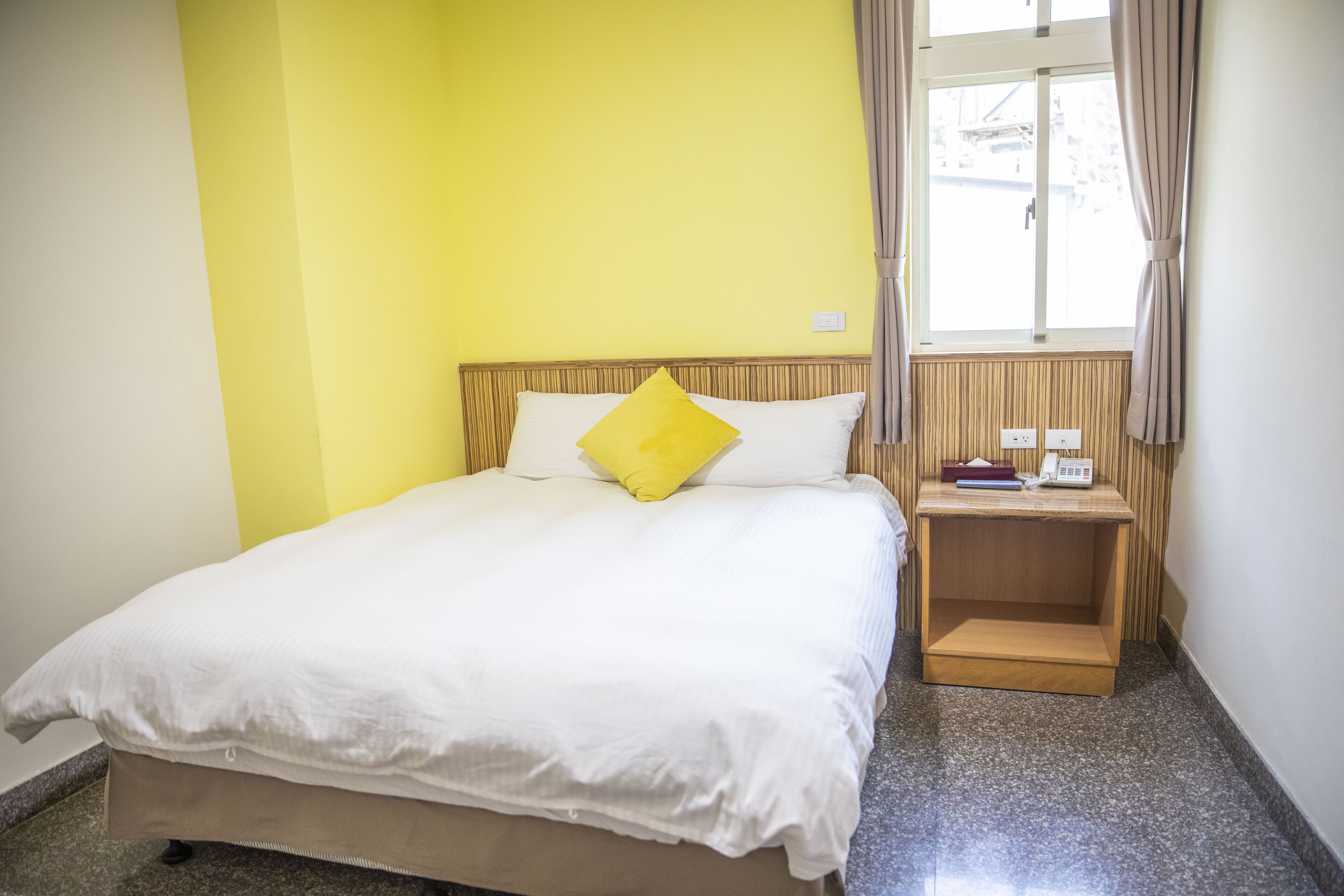 A bed with white bedding and a yellow pillow, a wooden nightstand with a phone and books, and a window with curtains.